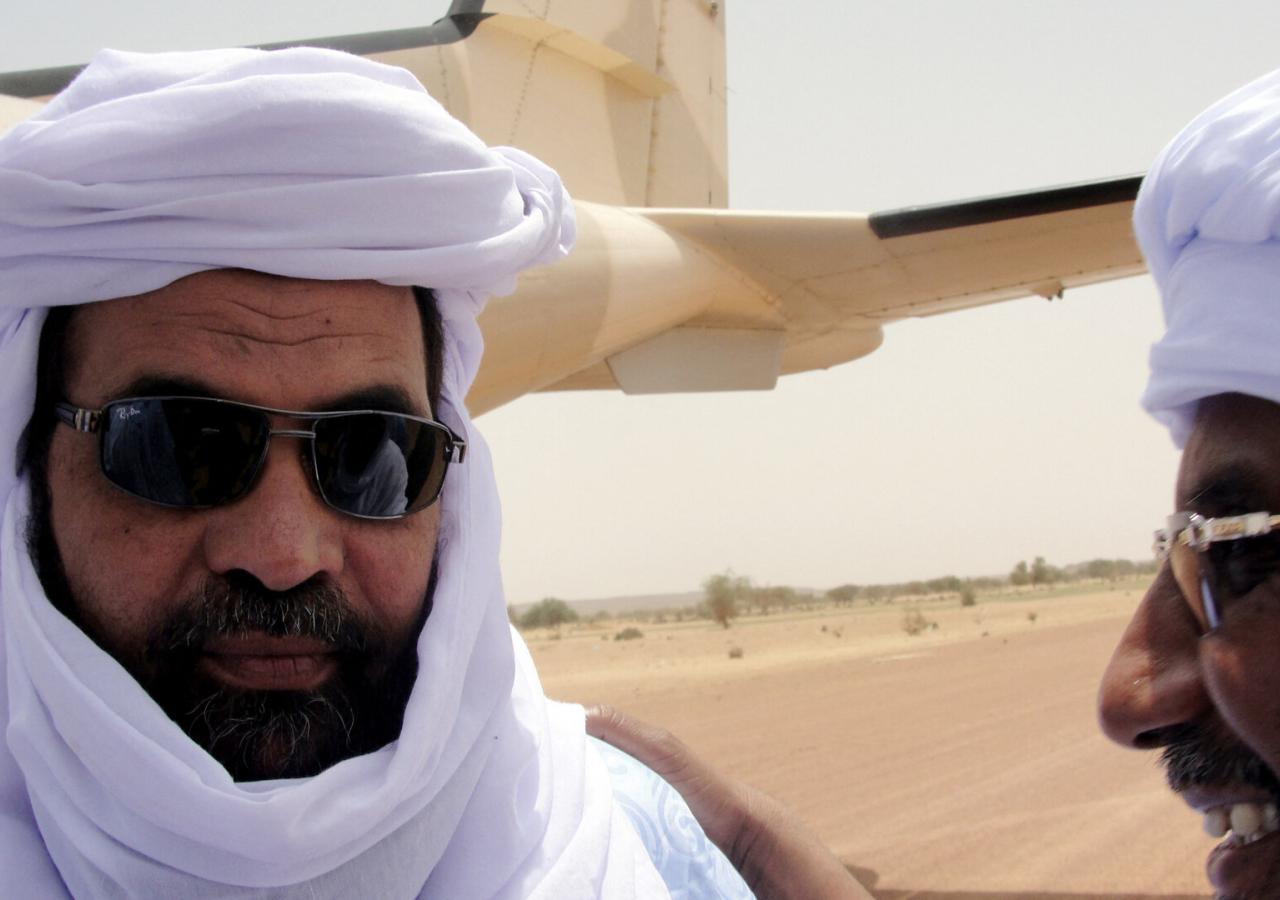 Iyad Ag Ghali in weißen Schleier mit Bart und Sonnenbrille. Er steht vor einem Flugzeug