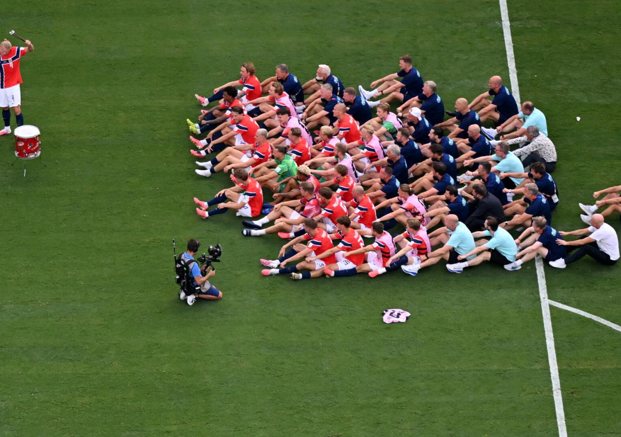 Die Norwegische Nationalmannschaft sitzt auf dem Fußballfeld und macht Ruderbewegungen zum Trommeltakt von E. Haaland