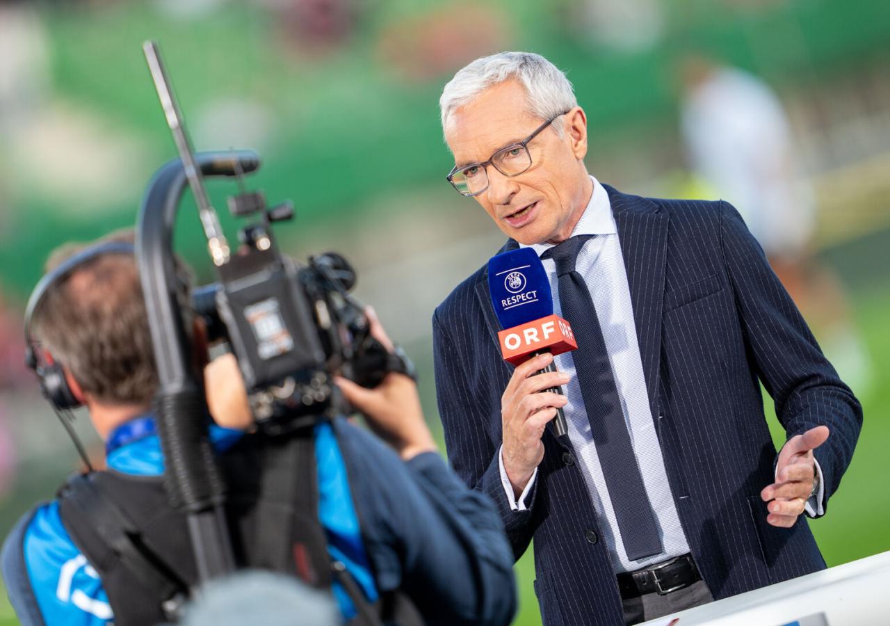 Moderator Rainer Pariasek steht mit Mikrofon vor einem Kameramann und macht eine Moderation