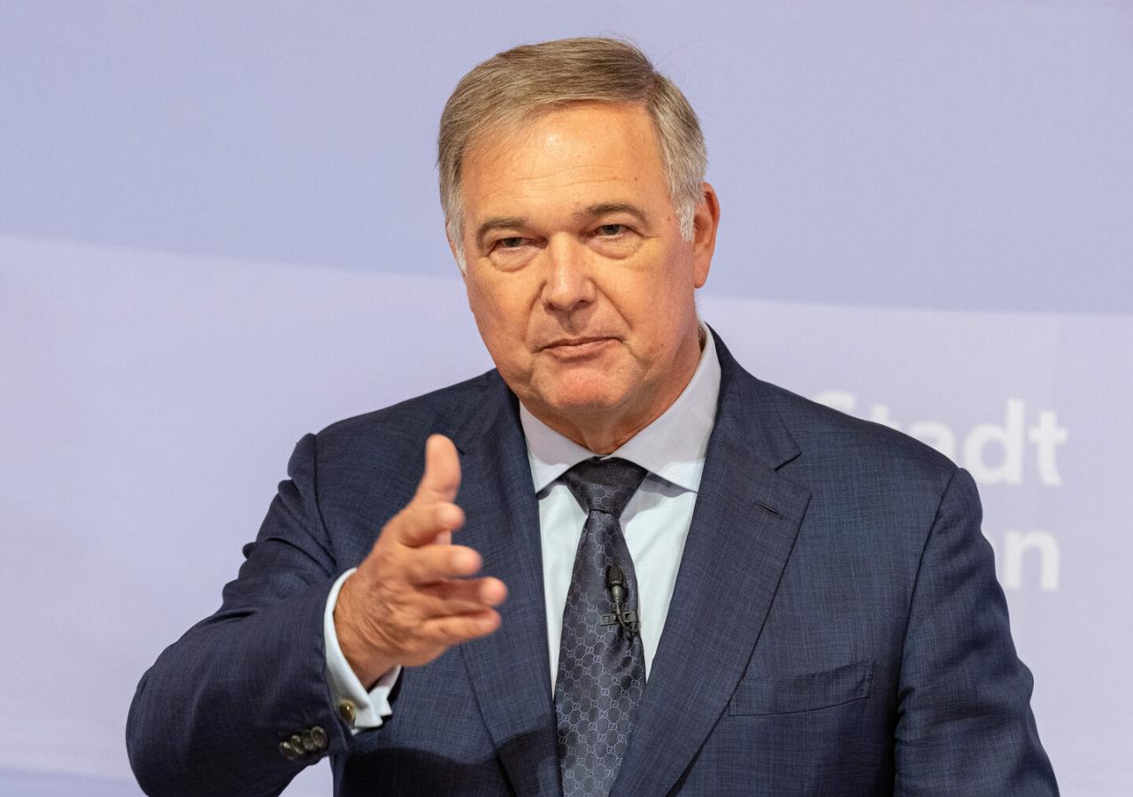Ein Mann im Anzug spricht mit ausgestreckter Hand vor hellviolettem Hintergrund.