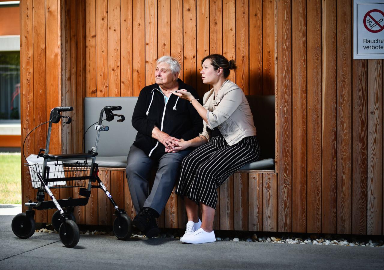Eine junge Frau spricht tröstend mit einer älteren Frau, die mit Rollator auf einer Holzbank sitzt.