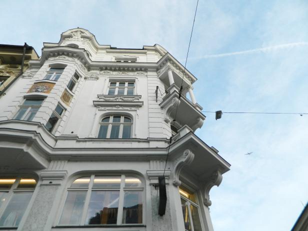 Ein weißes, reich verziertes Gebäude mit großen Fenstern und Balkonen vor blauem Himmel, im Hintergrund fliegt ein Flugzeug.