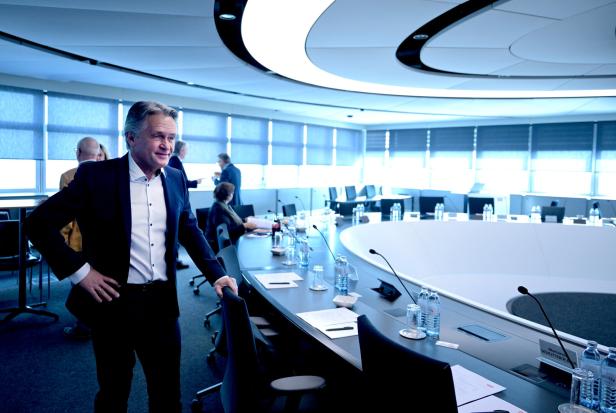 Ein Mann im Anzug, es ist Peter Westenthaler, steht in einem Konferenzraum mit ovalem Tisch und Mikrofonen.