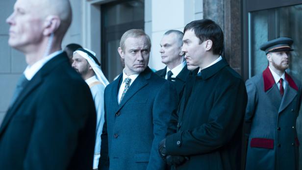 Jude Law als Wladimir Putin (Mitte), Paul Dano als sein Einflüsterer (Zweiter von rechts) Männer in dunklen Anzügen und Uniformen bei einer Art Empfang, zwei davon tauschen Blicke aus.