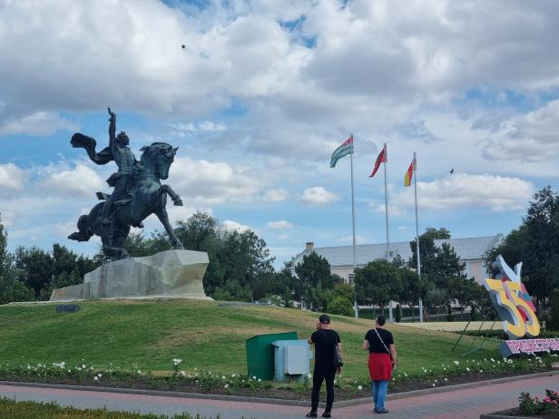 Zwei Personen betrachten eine Statue eines Reiters in der Mitte einer Grünfläche.