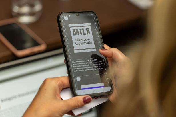 Eine Person hält ein Smartphone mit der App „MILA Mitmach-Supermarkt“ in der Hand.
