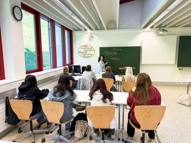 Mehrere Schülerinnen und Schüler sitzen in einem Klassenraum und hören einer Lehrerin zu, die an der Tafel steht und erklärt.