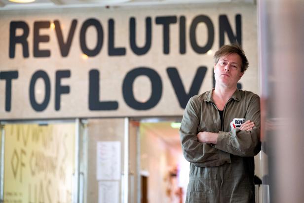 Mann im Overall mit verschränkten Armen, vor einem Transparent, auf dem "Revolution of Love" zu lesen ist.