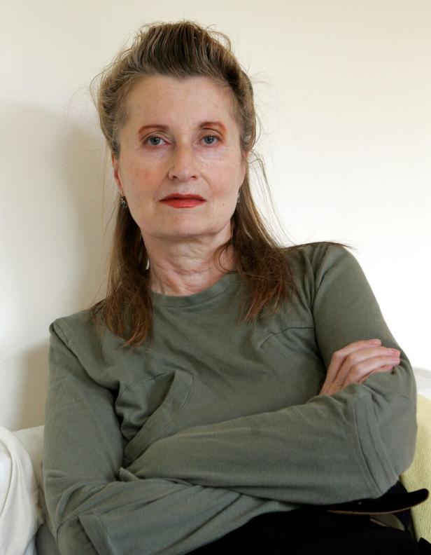 Frau sitzt mit ernstem Gesicht und verschränkten Armen vor einer weißen Wand.