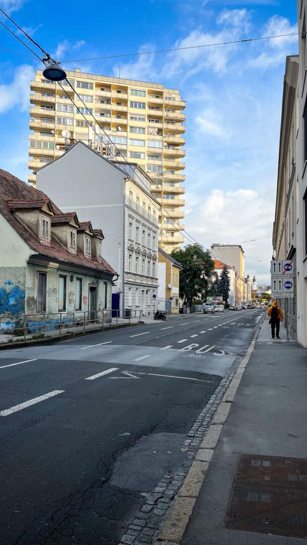 Im Bezirk Gries suchen einige Wohnungen neue Mieterinnen. An einer langen Straße im Grazer Wohnbezirk Gries stehen unterschiedliche Wohnhäuser: Ein heruntergekommenes kleines Häuschen, ein renovierter Altbau und ein gelbes Wohnhochhaus mit vielen Balkonen stehen hier. Der Himmel ist blau und leicht bewölkt. Bis auf einen einzelnen Fußgänger mit gelber Jacke ist die Straße leer.