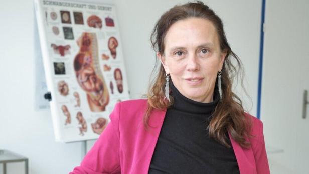 Mag.a(FH) Beate Kayer Beate Kayer hat braune Haare und trägt einen pinken Blazer. Im Hintergrund eine medizinische Grafik.