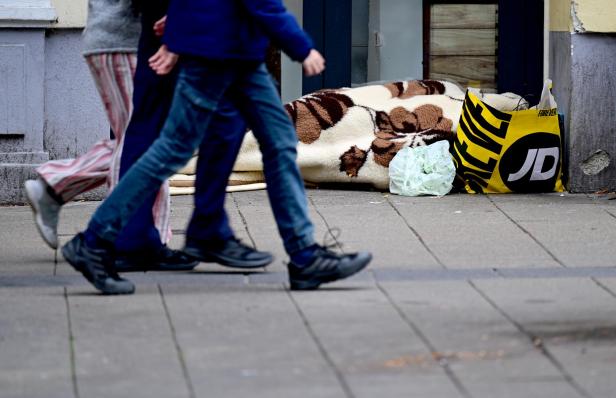 Mehrere Personen gehen an einer schlafenden Person vorbei, die unter einer Decke auf dem Gehweg liegt, daneben Taschen mit Habseligkeiten.
