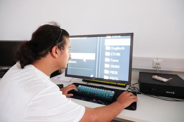 Eine Person sitzt an einem Schreibtisch vor einem großen Monitor und öffnet digitale Ordner über Tastatur und Maus.