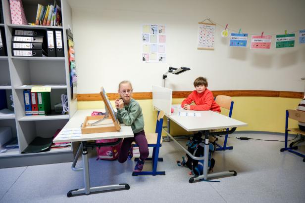 In einem hellen Klassenzimmer sitzen zwei Kinder an nebeneinander stehenden Tischen. Vor ihnen liegen bunte Lernkarten und spezielle Arbeitsmaterialien; eines der Kinder nutzt eine schräg aufgestellte Lern- oder Lesetafel. Hinter ihnen hängen farbige Unterrichtsposter an der Wand.