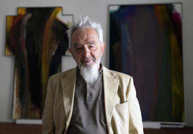 Der Künstler Arnulf Rainer blickt ernst in die Kamera. Hinter ihm zwei seiner Übermalungen.
