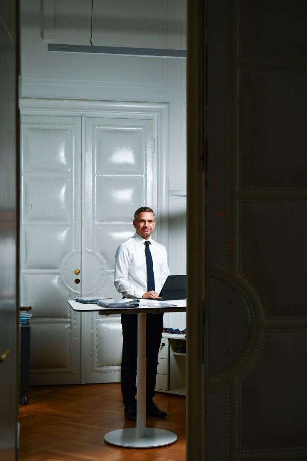 Ein Mann im Hemd und Krawatte, Wolfgang Hattmannsdorfer, steht an einem Stehtisch mit Laptop und Unterlagen in einem eleganten Büro.