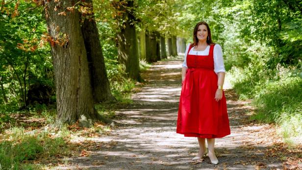 Barbara Karlich steht in rotem Dirndl-Kleid auf einem Waldweg