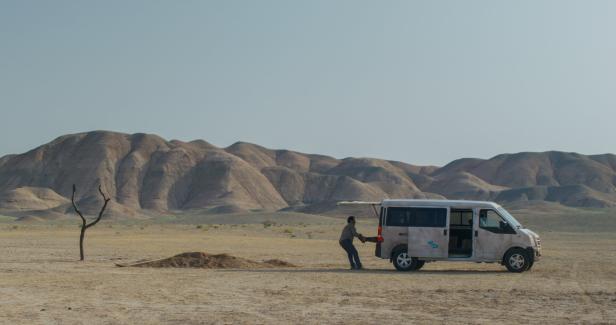 Ein weißer Kombi steht, aus der Ferne gesehen, in der Wüste: Ein Mann zieht einen anderen an den Füßen aus dem Kofferraum.
