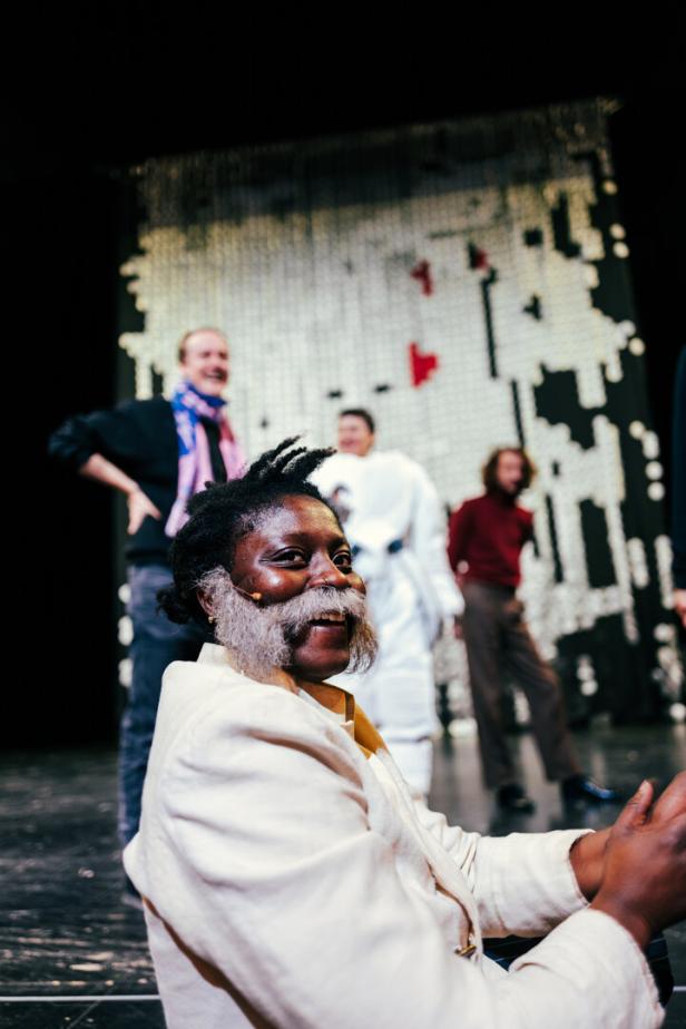 Die Schauspielerin Nancy Mensah-Offei lacht mit Backenbart im Bildvordergrund, hinter ihr auf der Bühne: Regisseur Jan-Christoph Gockel, Darstellerin Alicia Aumüller und ihr Kollege Bernardo Arias Porras
