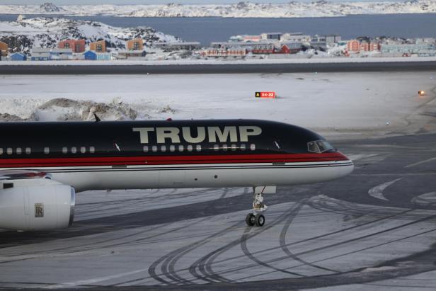 Ein Flugzeug mit der Aufschrift "Trump" rollt über eine verschneite Landebahn. Im Hintergrund sind Häuser, Meer und Berge von Grönland zu sehen.