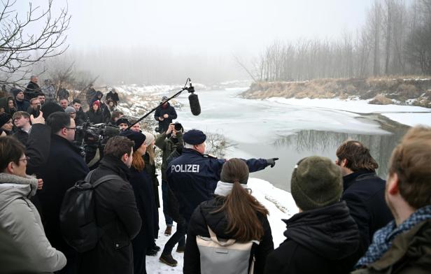 Eine Gruppe von Menschen steht am verschneiten Flussufer, ein Polizist zeigt auf das teilweise zugefrorene Wasser.