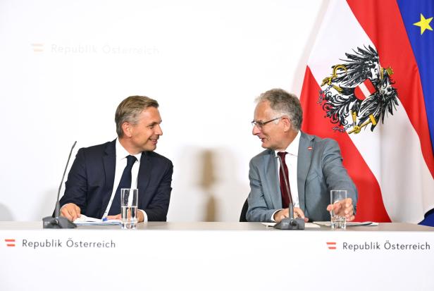 Zwei Männer - die Minister Hattmannsdorfer und Marterbauer - in Anzügen sitzen an einem Tisch vor der österreichischen und der EU-Flagge.