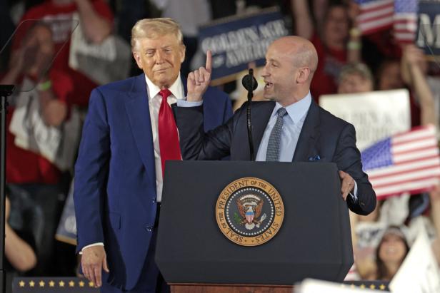 Miller spricht am Podium, Trumps steht hinter ihm, im Hintergrund Fans mit US-Flaggen