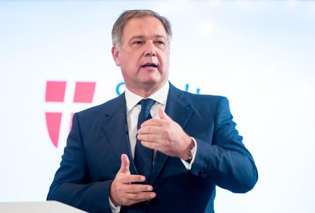 Ein Mann im Anzug spricht gestikulierend vor einem Rednerpult mit Wappen im Hintergrund.