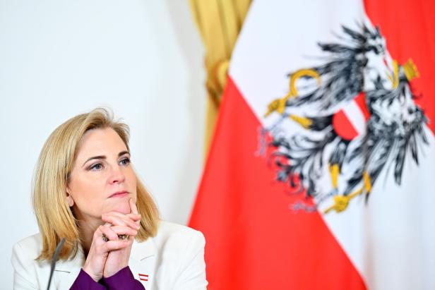 Eine Frau mit blondem Haar sitzt nachdenklich vor einer österreichischen Flagge mit Bundesadler.