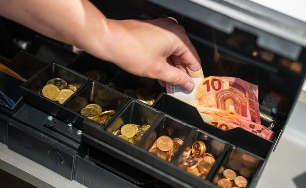 Eine Hand legt einen Zehn-Euro-Schein in eine geöffnete Ladenkasse mit Münzfächern.
