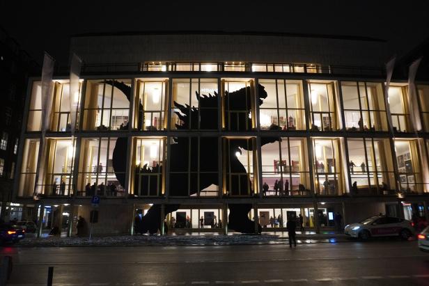 Die Silhouette eines gigantischen Sauriers, nachts an der gläsernen Fassade eines von innen erleuchteten Hauses 