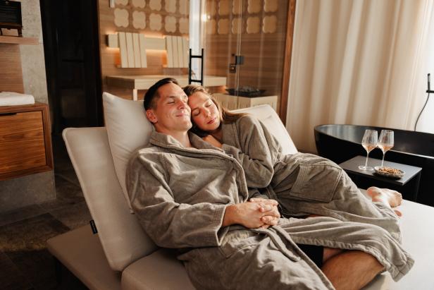 Paar in Bademänteln entspannt auf einer Liege in einem modernen Wellnessbereich mit Badewanne und Weingläsern im Hintergrund
