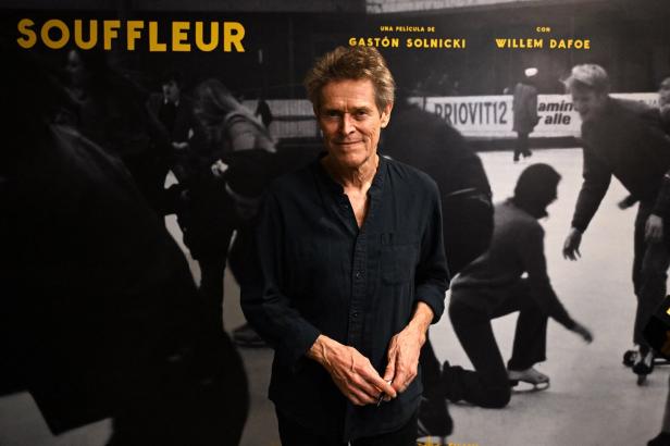 Willem Dafoe lächelnd vor einem Plakat zu seinem jüngsten Film, "The Souffleur" 
