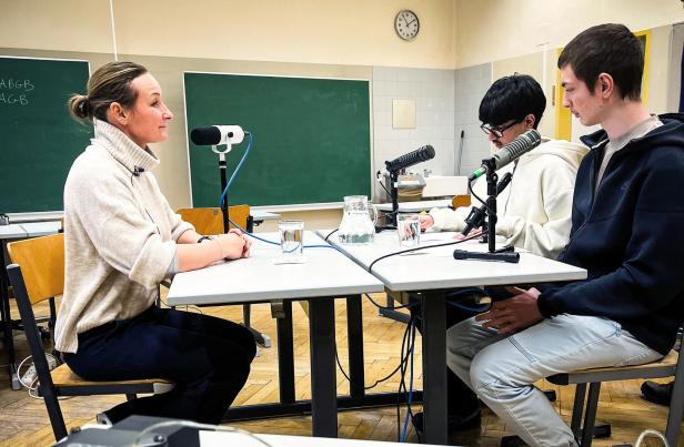 Drei Personen sitzen mit Mikrofonen an Tischen in einem Klassenzimmer und nehmen einen Podcast auf.