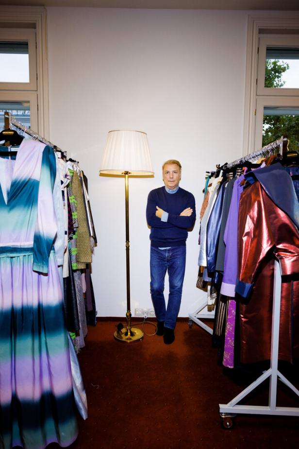 Modedesigner Atil Kutoglu steht zwischen zwei Garderobenständern mit bunten Kleidern