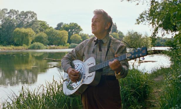 Al Cook alias Alois Koch mit silberner akustischer Gitarre in einer Au-Landschaft