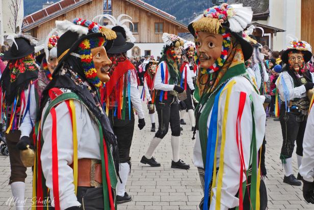 Mehrere Menschen tragen bunte Trachten, Masken und Hüte mit Federn bei einem traditionellen Umzug im Freien.