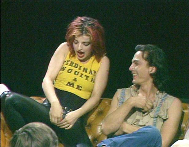 Nina Hagen fasst sich, in gelbem T-Shirt und schwarzer Lederhose, in einem Fernsehstudio demonstrativ mit beiden Händen in den Schritt; ihr damaliger Partner Ferdinand Karmelk amüsiert sich neben ihr.