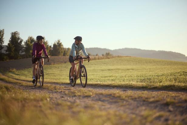 Zwei Personen fahren mit Fahrrädern auf einem Schotterweg durch eine grüne Landschaft bei Sonnenschein.