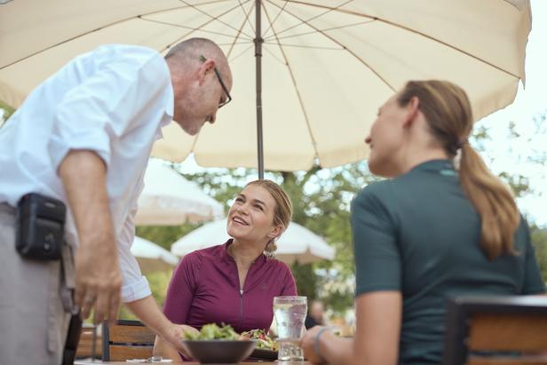 Ein Kellner bedient zwei Frauen an einem Tisch im Freien unter einem Sonnenschirm.