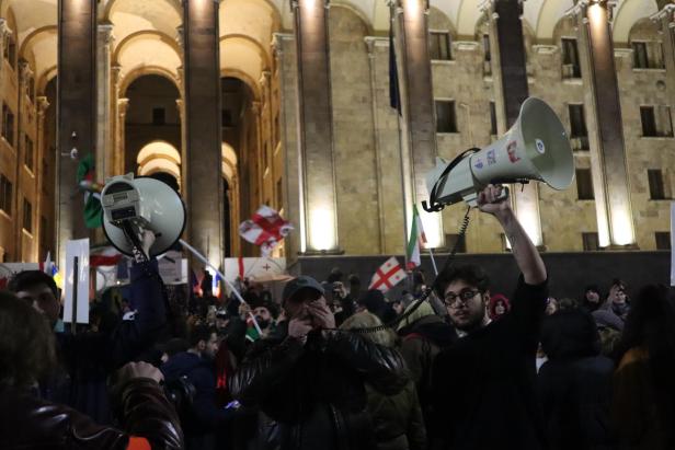 Jemand spricht in zwei Megafone vor dem georgischen Parlament (ein großes Gebäude aus hellem Vulkanstein, das beleuchtet wird)