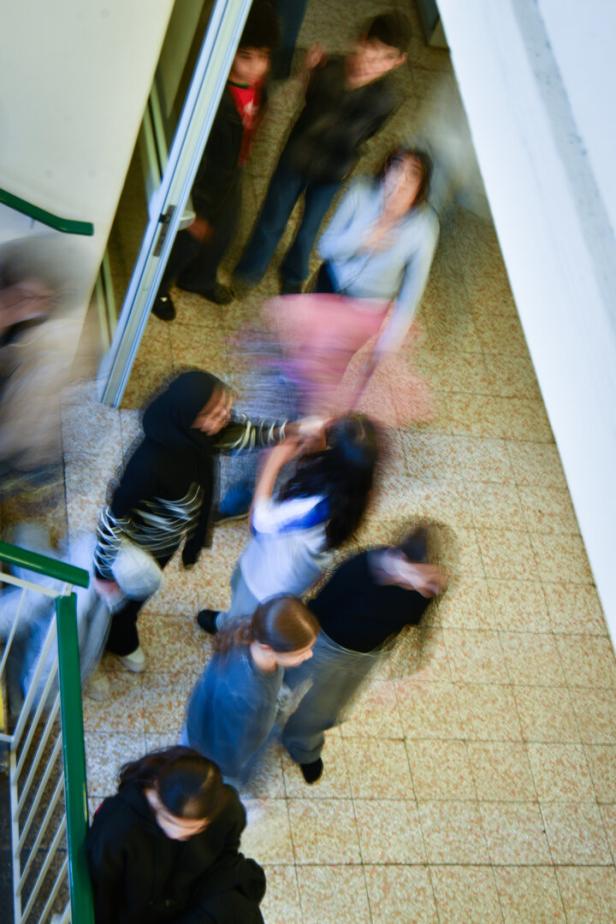 Mehrere Jugendliche bewegen sich verschwommen durch einen Flur mit Fliesenboden und Treppengeländer.