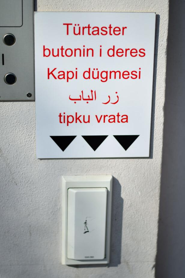 Mehrsprachiges Schild mit der Aufschrift „Türtaster“ und einem weißen Taster darunter an einer Wand.