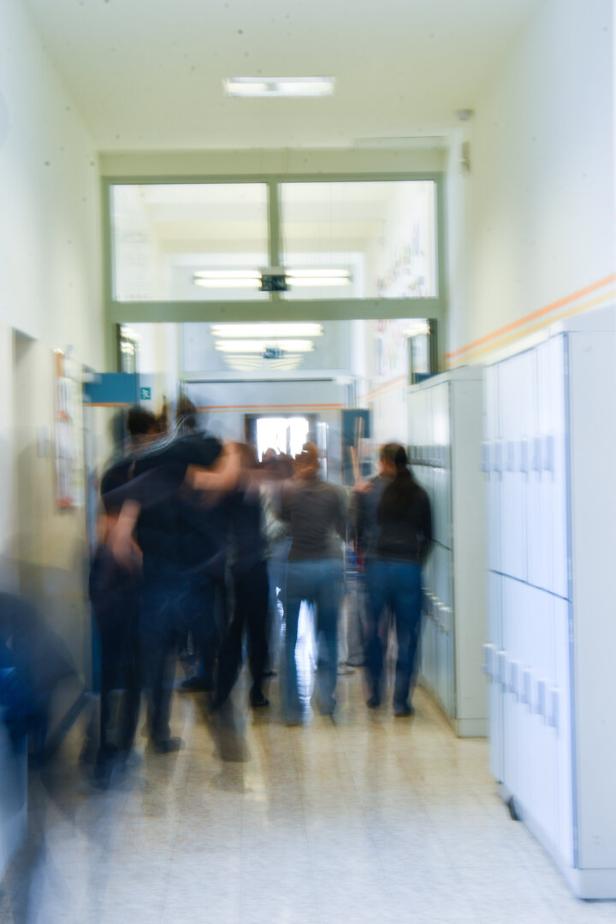 Mehrere unscharfe Personen gehen durch einen hellen Flur mit weißen Spinden an der Wand.