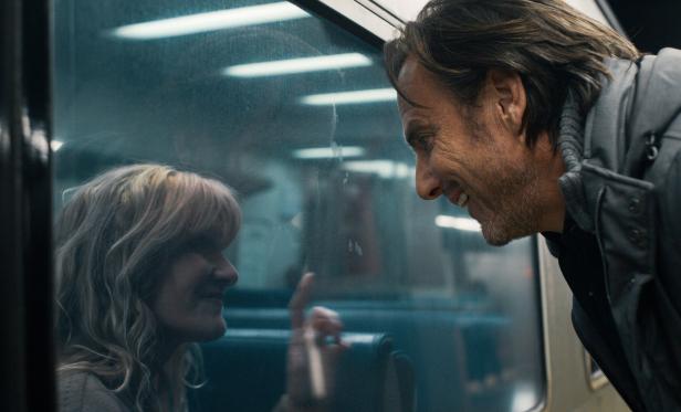 Will Arnett blickt grinsend auf Laura Dern durch die Fensterscheibe der New Yorker U-Bahn.