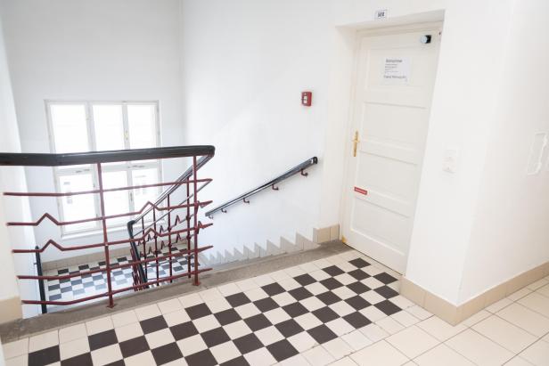 Helles Stiegenhaus mit rot-schwarzem Geländer und weißer Tür, daneben ein großes Fenster.