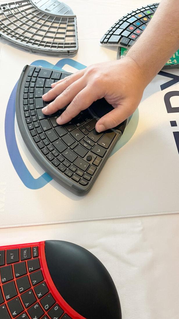 Eine Hand liegt auf einer ergonomisch geformten, halbkreisförmigen Tastatur mit mehreren ähnlichen Tastaturen im Hintergrund.