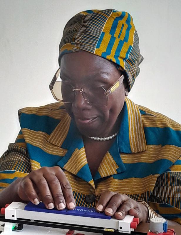 Eine Frau mit Brille und traditioneller Kleidung aus Simbabwe liest mit den Fingern an einer Brailleschrift-Tafel.