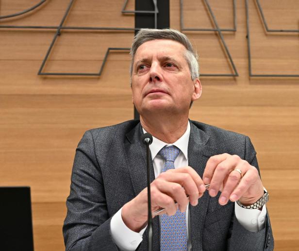 Ein Mann im Anzug sitzt an einem Tisch vor einem Mikrofon und blickt nachdenklich zur Seite.