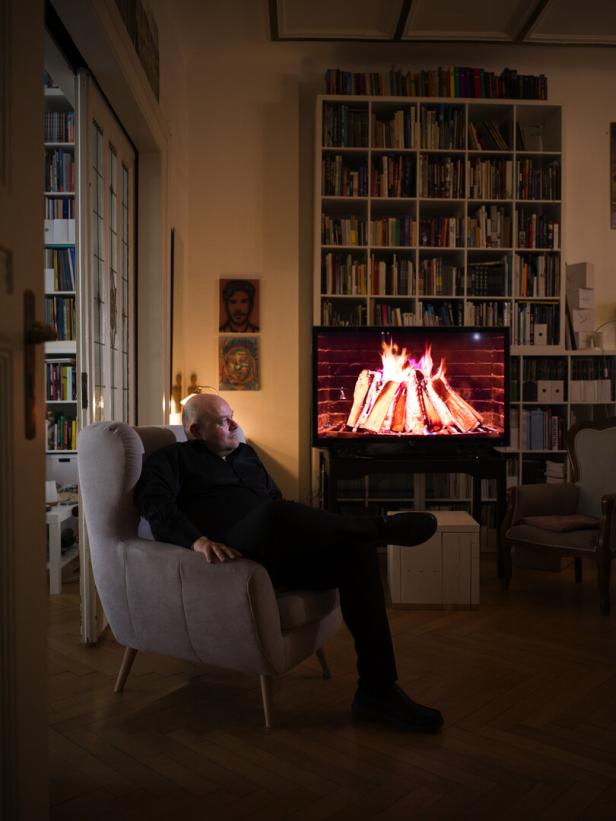 Filmemacher Michal Kosakowski sitzt in einem Lehnstuhl in einer Wohnung, im Hintergrund ein Bücherregal und ein Bildschirm, auf dem ein Kaminfeuer zu sehen ist